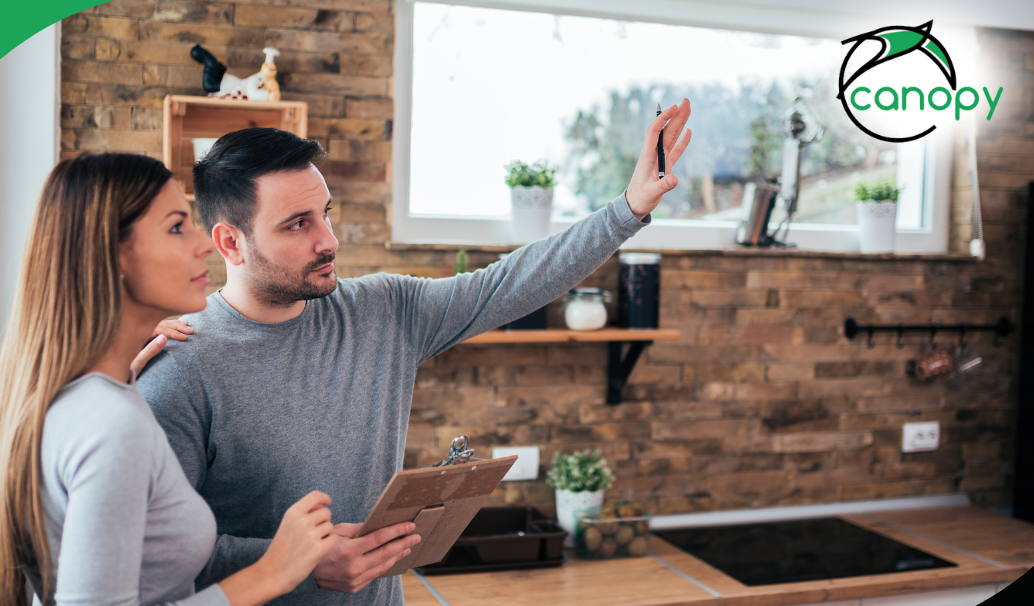A home inspector explaining details to a client, both looking at the inspection area, with a clipboard in hand.