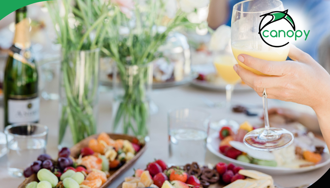 Close-up of a hand holding a glass of orange juice with a festive brunch spread in the background.
