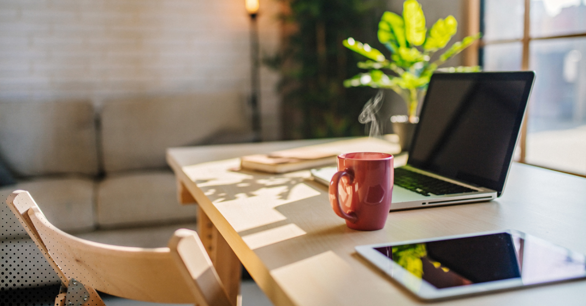A cozy workspace with a steaming coffee cup, a laptop, and an iPad on a wooden table, surrounded by plants in a well-lit room.